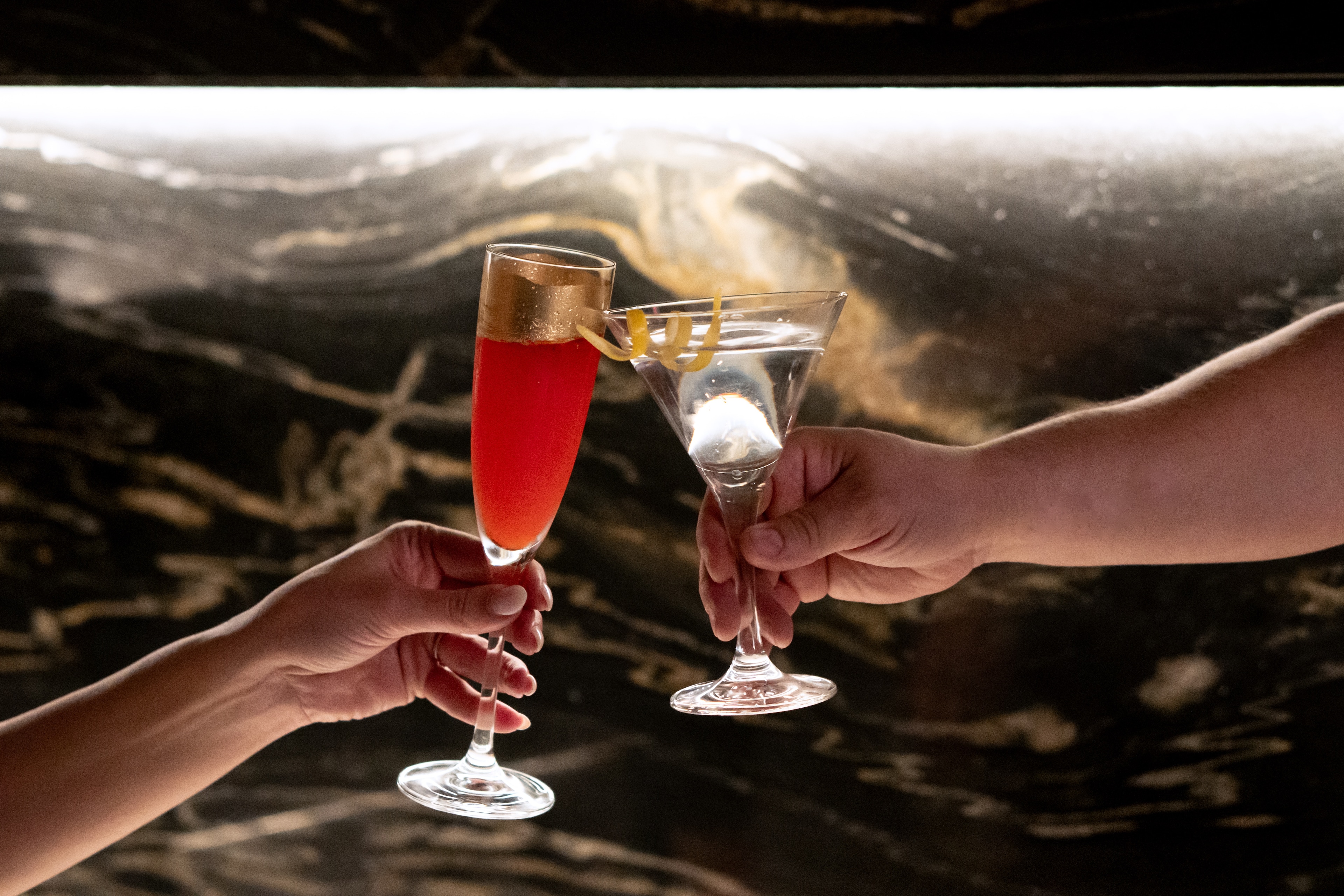 Two hands holding up cocktails in front of a marble background