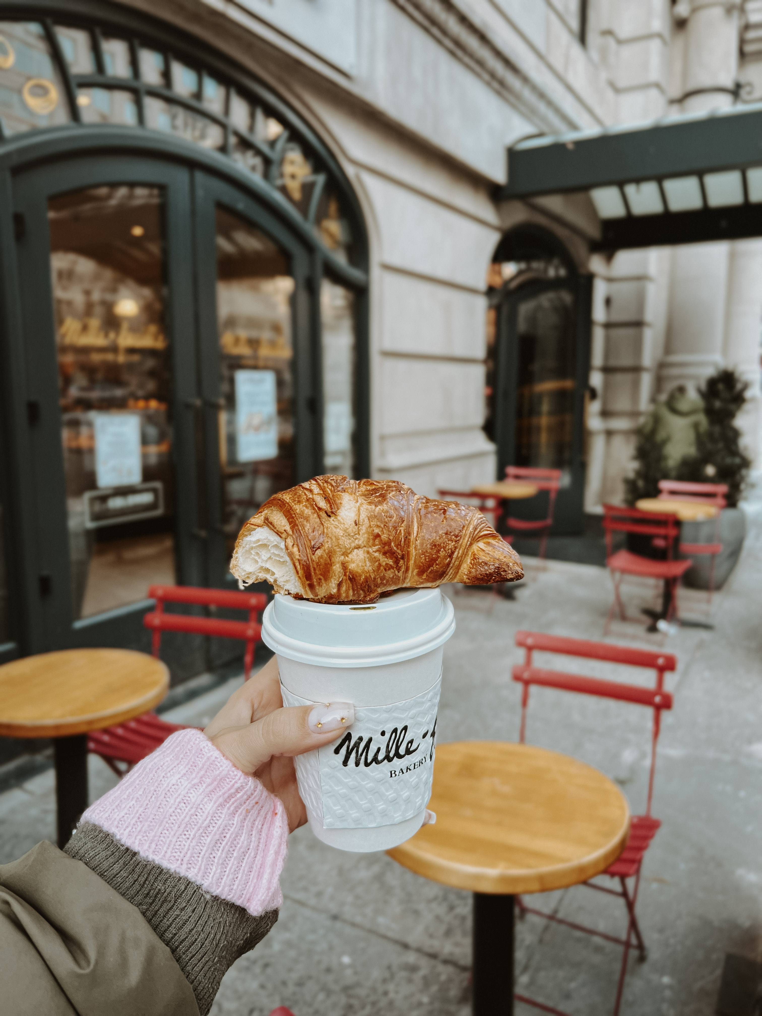 A hand holding a coffee cup with a croissant on top, outside a bakery.