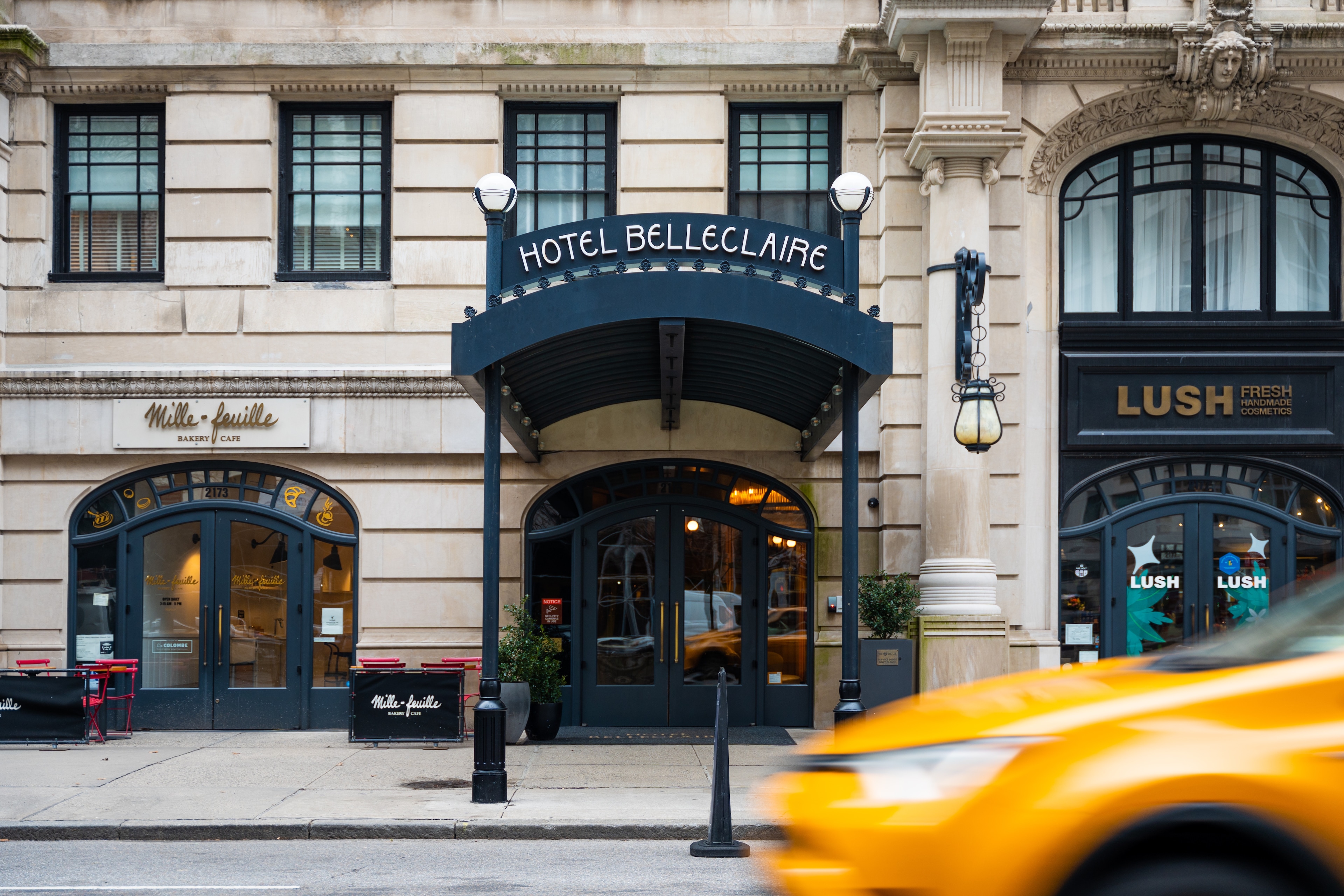 A photograph of the Hotel Belleclaire's exterior, showing its stone facade, black awning with the hotel's name, and storefronts including a Lush cosmetics store and a bakery cafe.