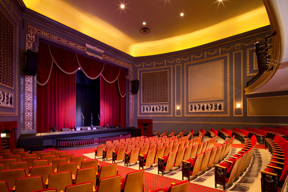 A luxurious meeting room with a stage, red curtains, gold molding, and theater seating.