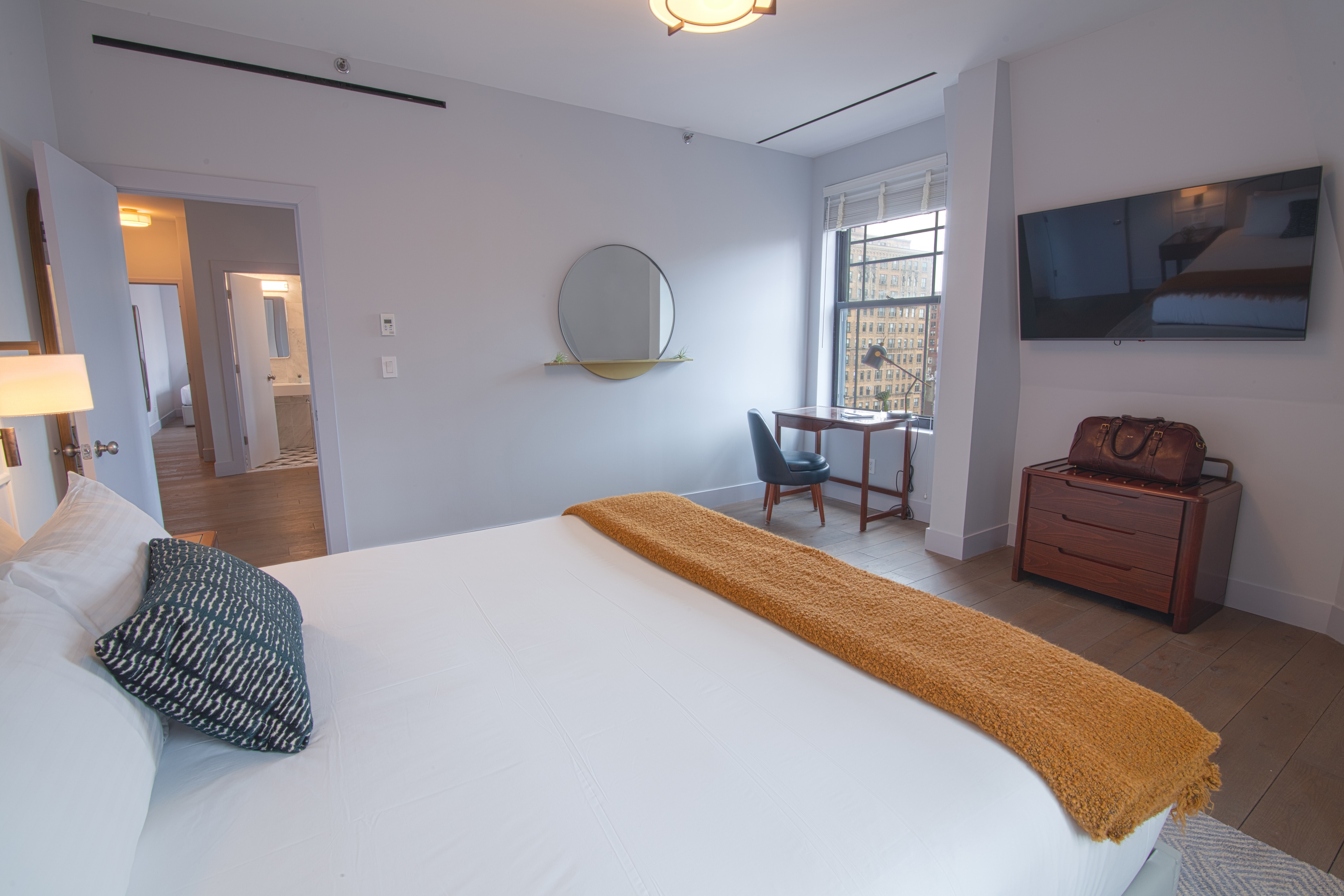 A hotel room with a large bed, a TV on the wall, a desk and chair, and a window with a city view.