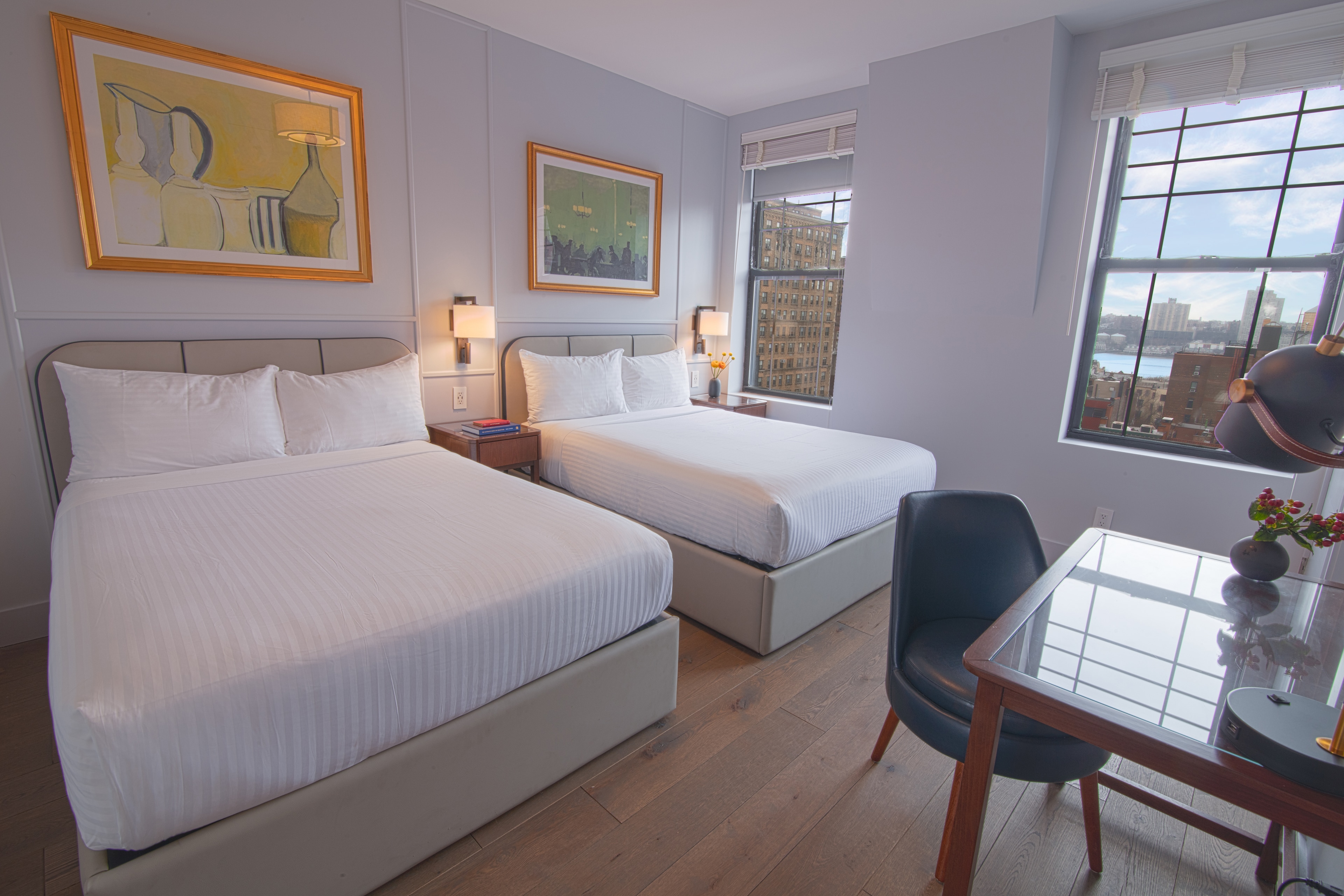 A hotel room with two beds, a desk, and a window showing a city view.