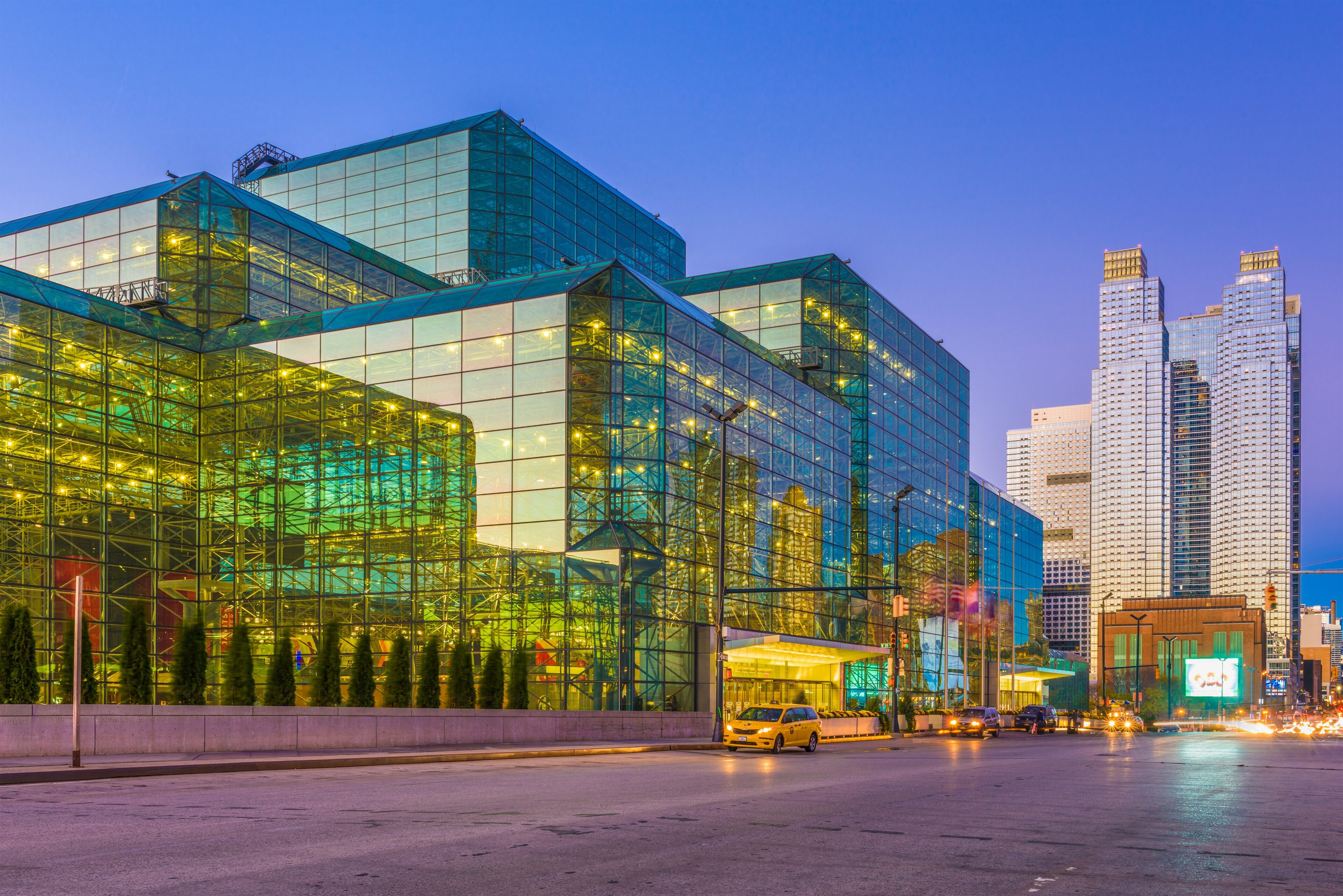 A large, cube-shaped building with a glass facade lit up at dusk or dawn with cars parked in front.
