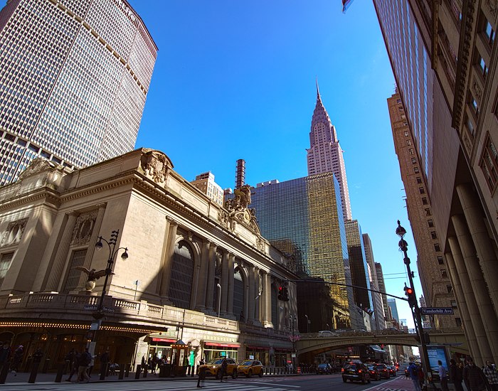 A wide-angle view of a large building on a city street corner with tall skyscrapers in the background under a clear blue sky.