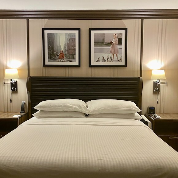 A wide view of a hotel room bed with a dark wood headboard, two nightstands with lamps, and framed artwork above the bed.