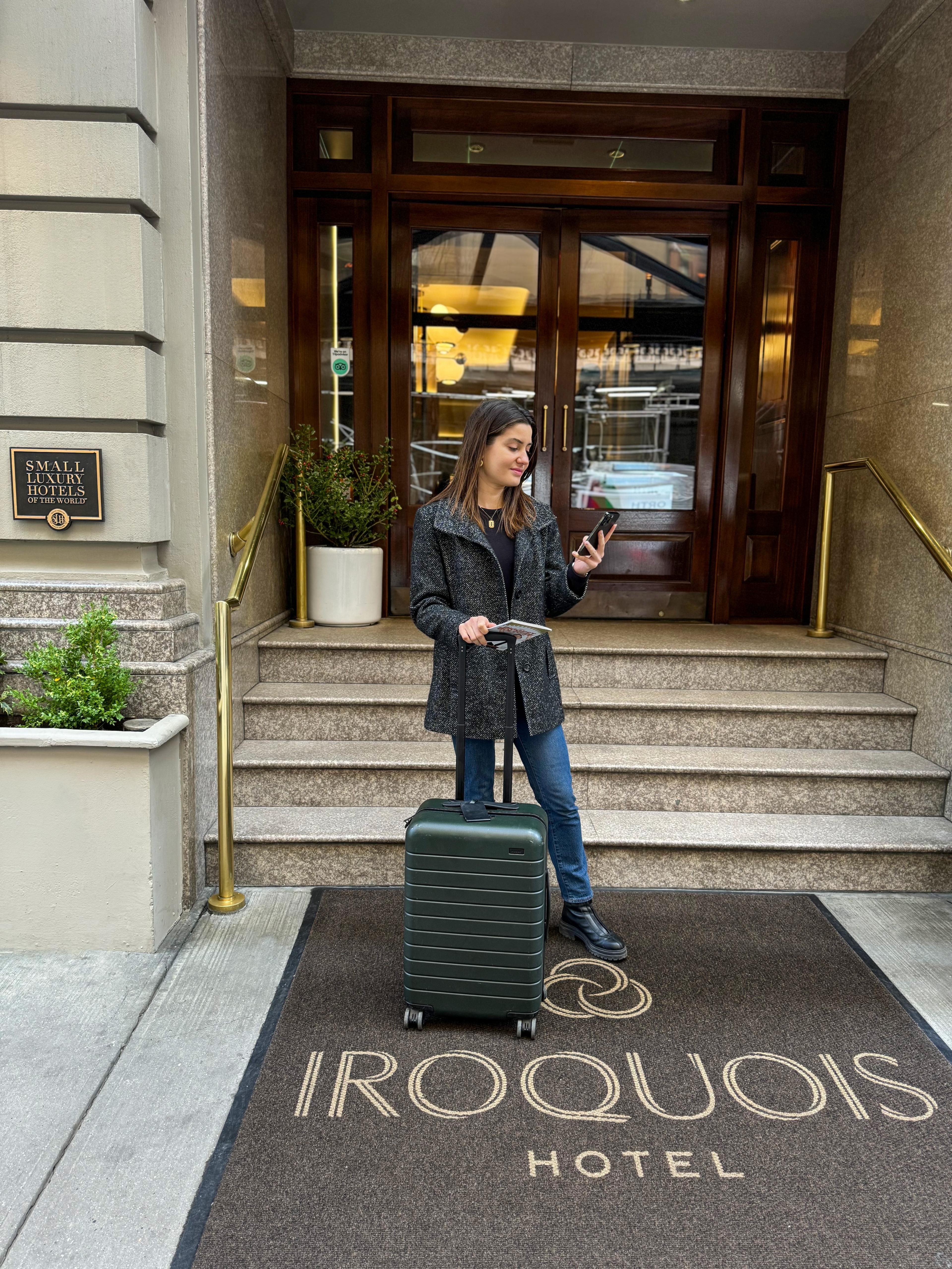 A woman stands on the steps of a hotel entrance with a suitcase and phone.
