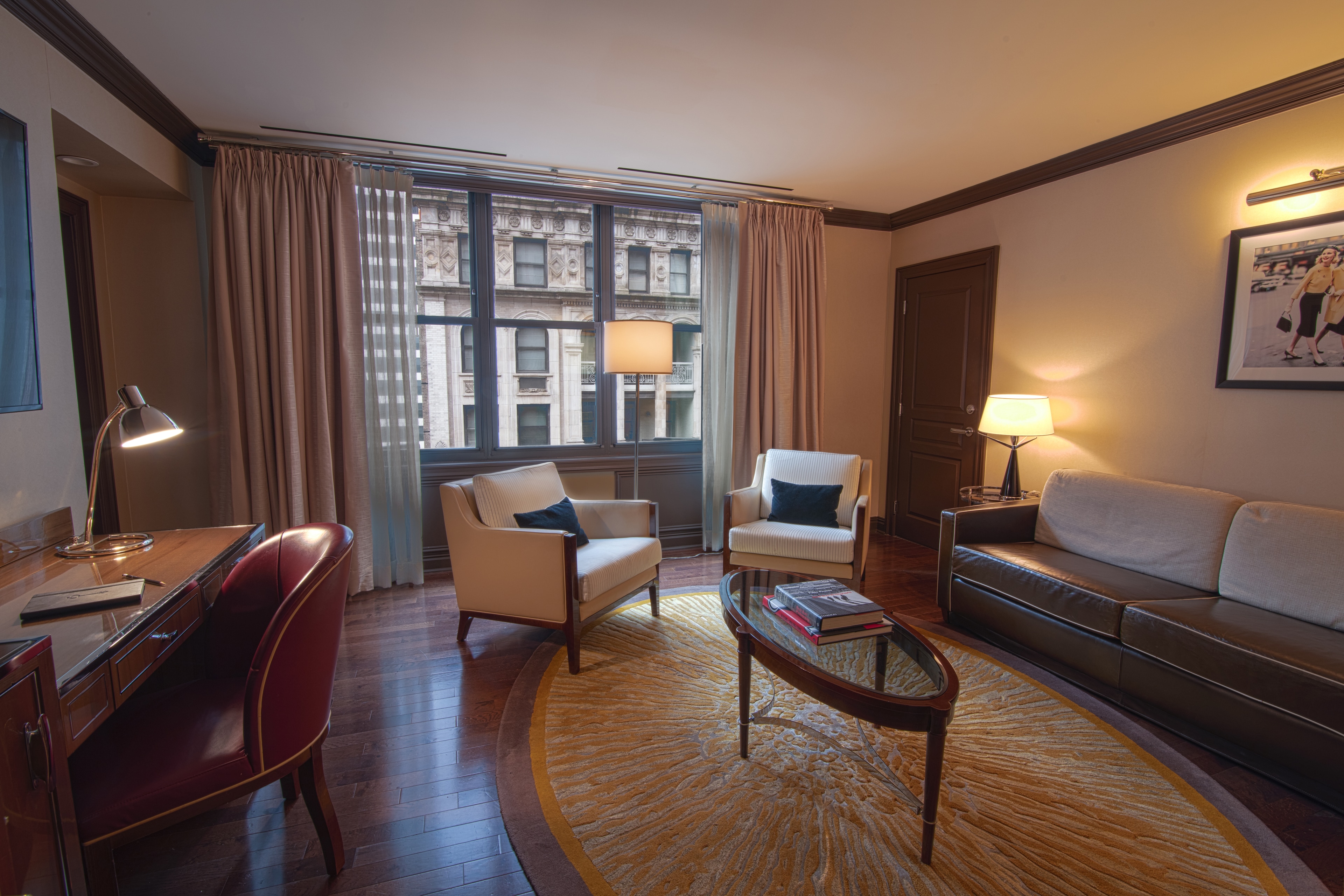A wide view of a hotel room with a sofa, two armchairs, a coffee table, a desk, and a large window showing the city view.