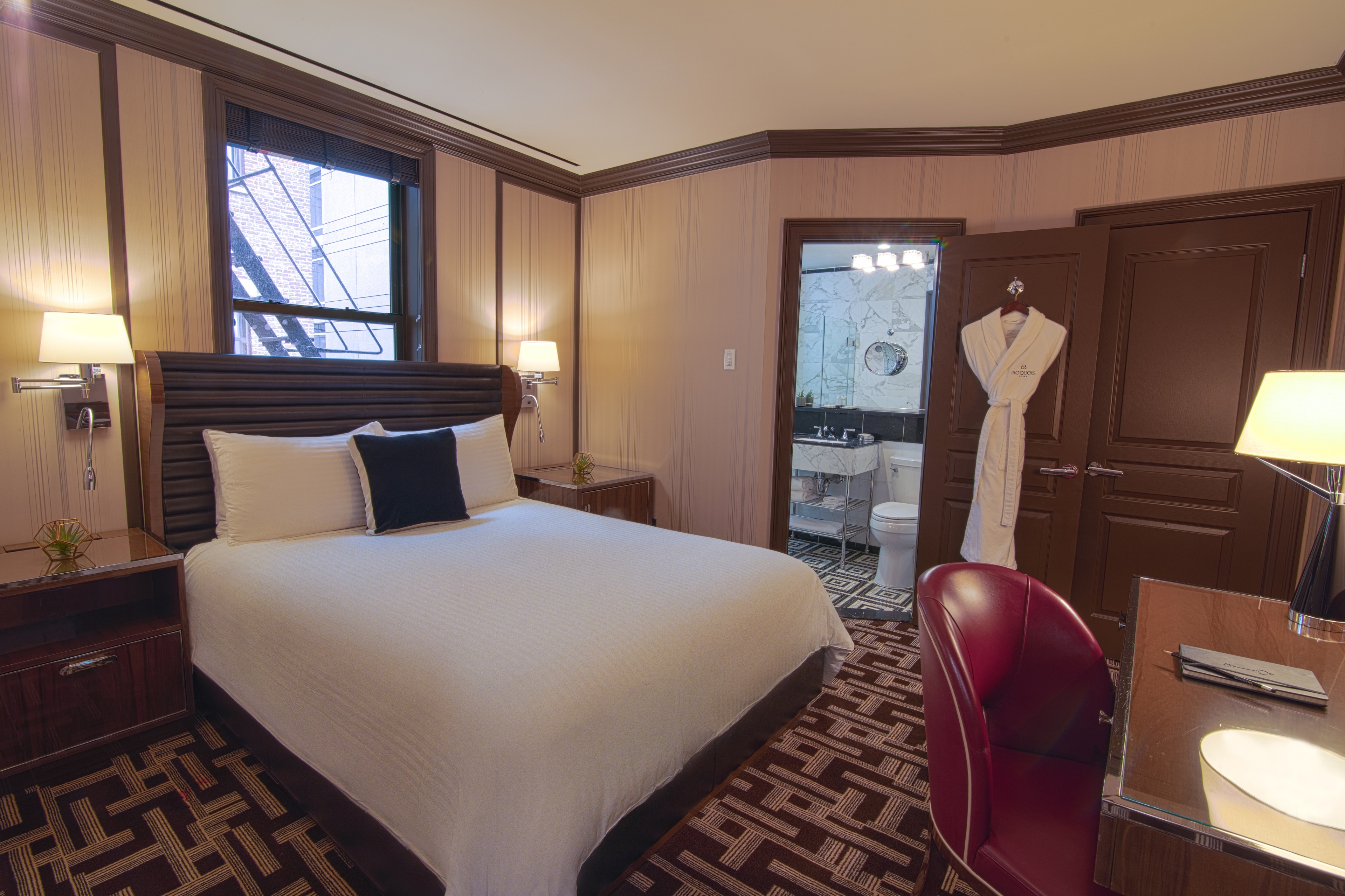 A hotel room with a bed, nightstand, lamp, window, and bathroom