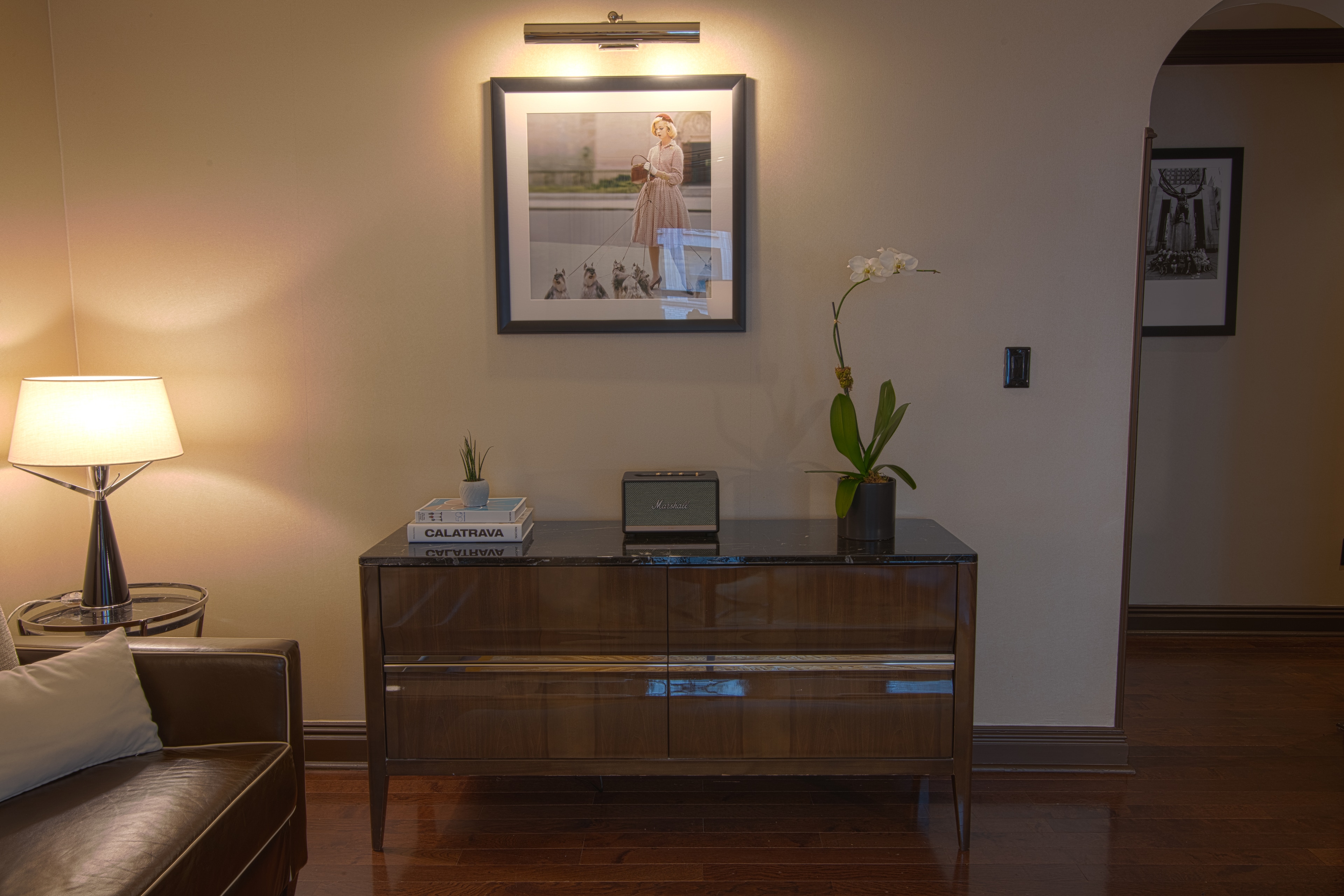 A hotel room with a brown leather couch, a wooden credenza with a lamp, books, radio, and plant on top, and a framed artwork on the wall.