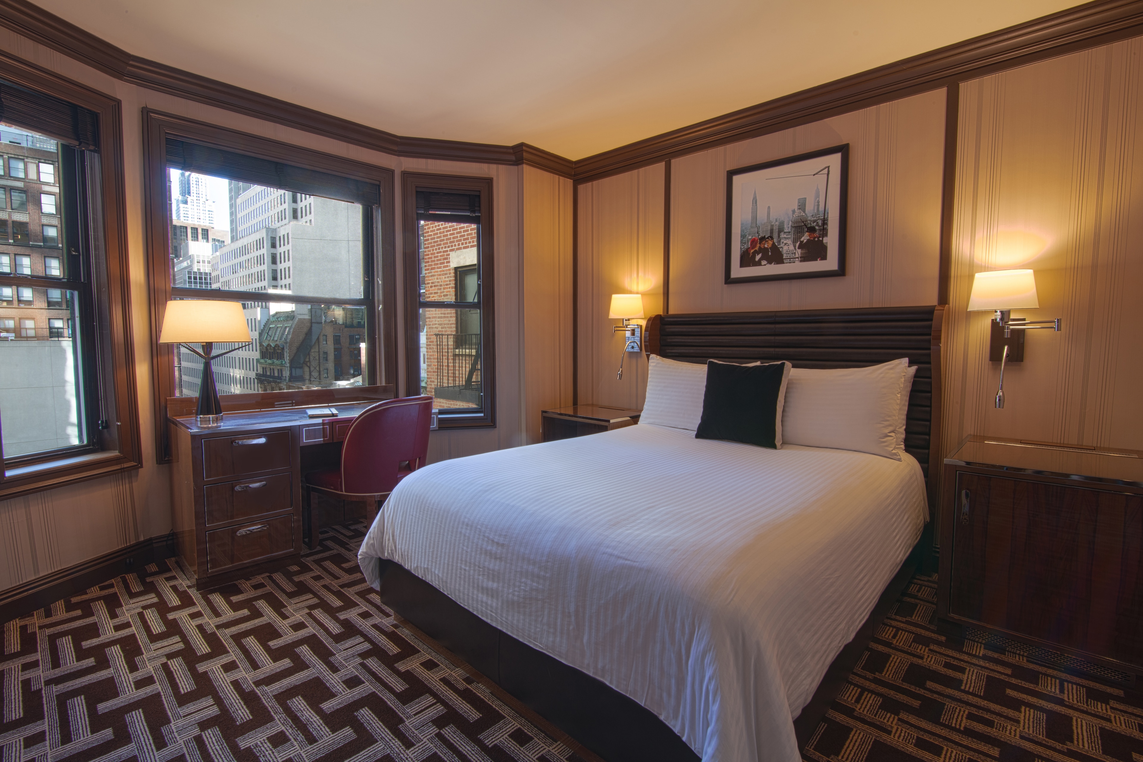 A hotel room with a large bed, desk, and window showing a city view.