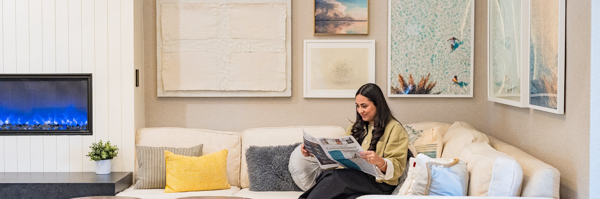 A woman sits on a beige sectional sofa reading a newspaper in a modern living room at Cambria Hotel Calabasas, featuring a gallery wall of framed art and a lit electric fireplace.