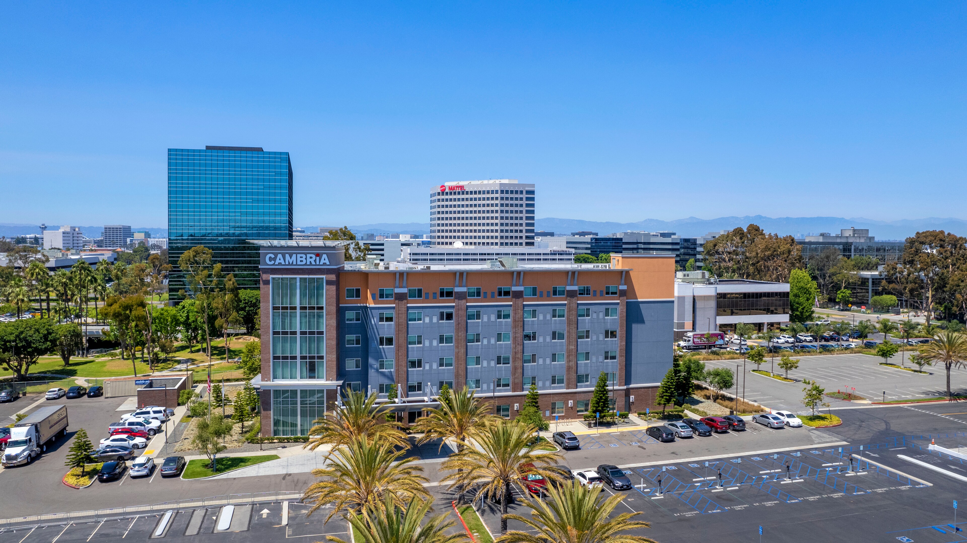 Aerial view of a modern hotel with a "Cambria" sign, surrounded by palm trees and parked cars