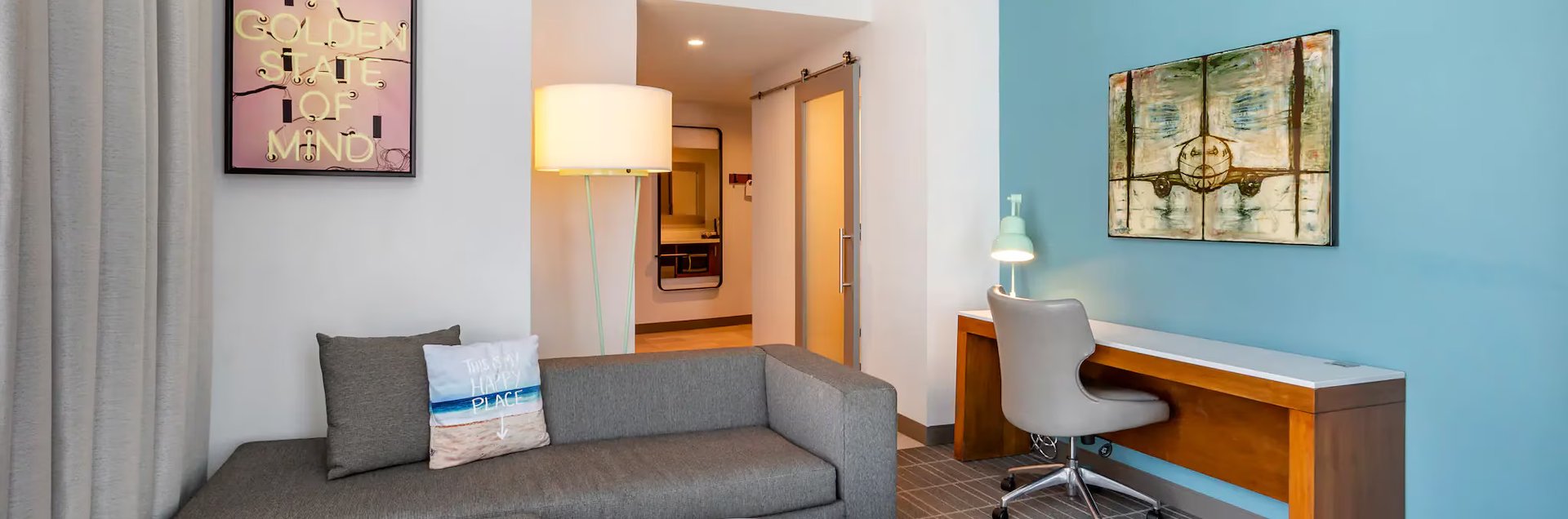 Modern hotel room with a blue accent wall, gray L-shaped sofa, and a sleek work desk