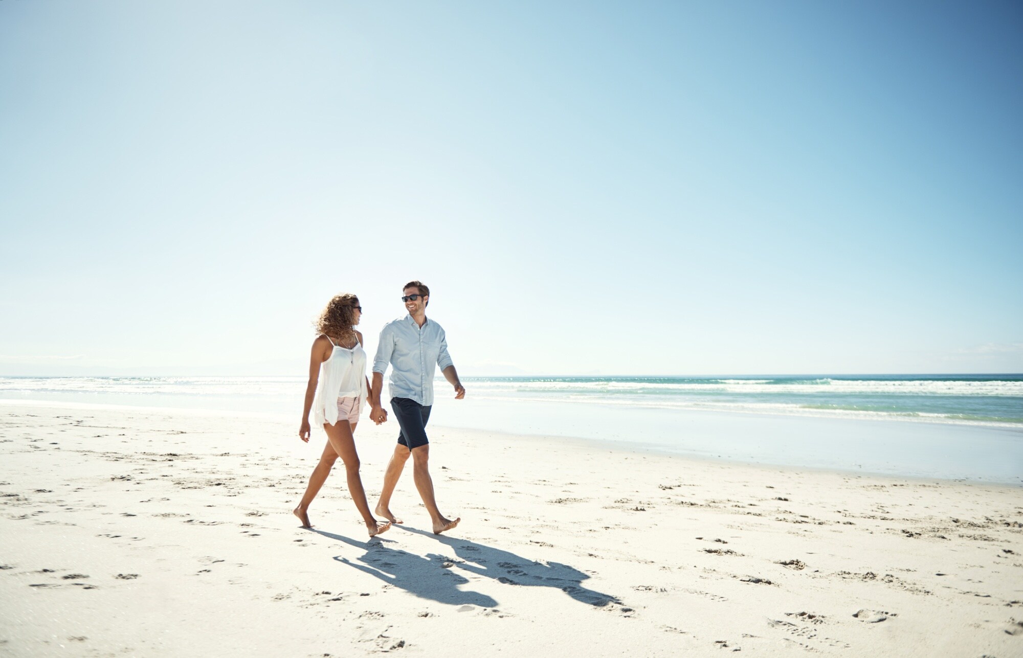 A man and woman walk hand in hand on a sunny beach, casting long shadows on the sand