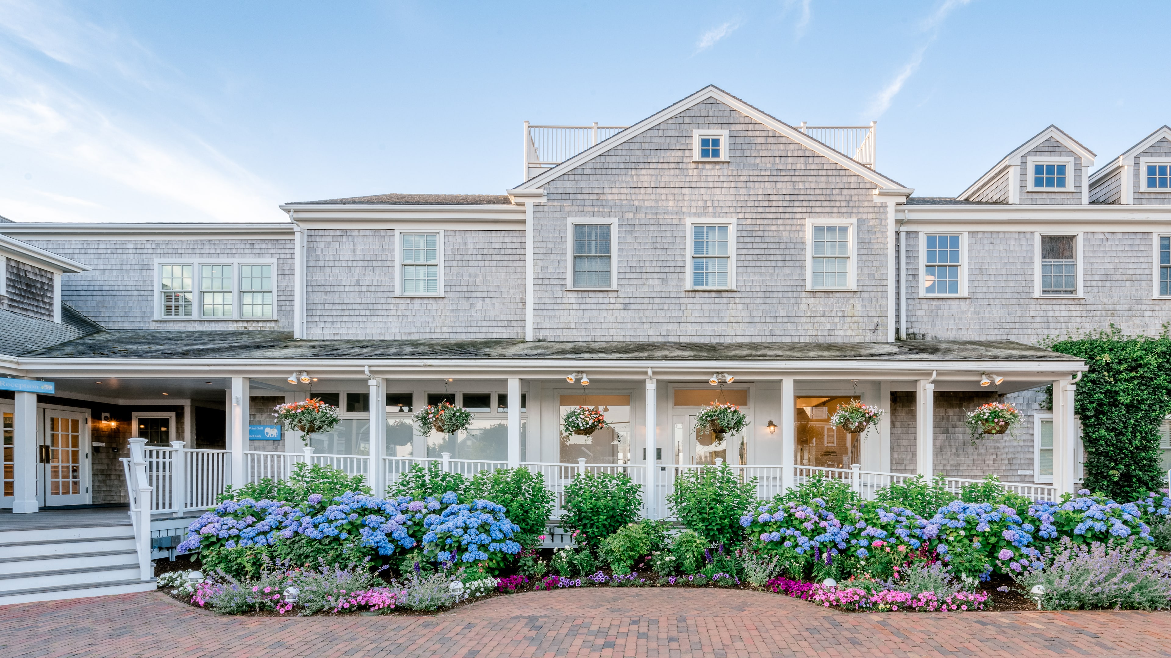 A large gray shingle building with a white porch and railing, surrounded by flowering plants and a brick driveway.