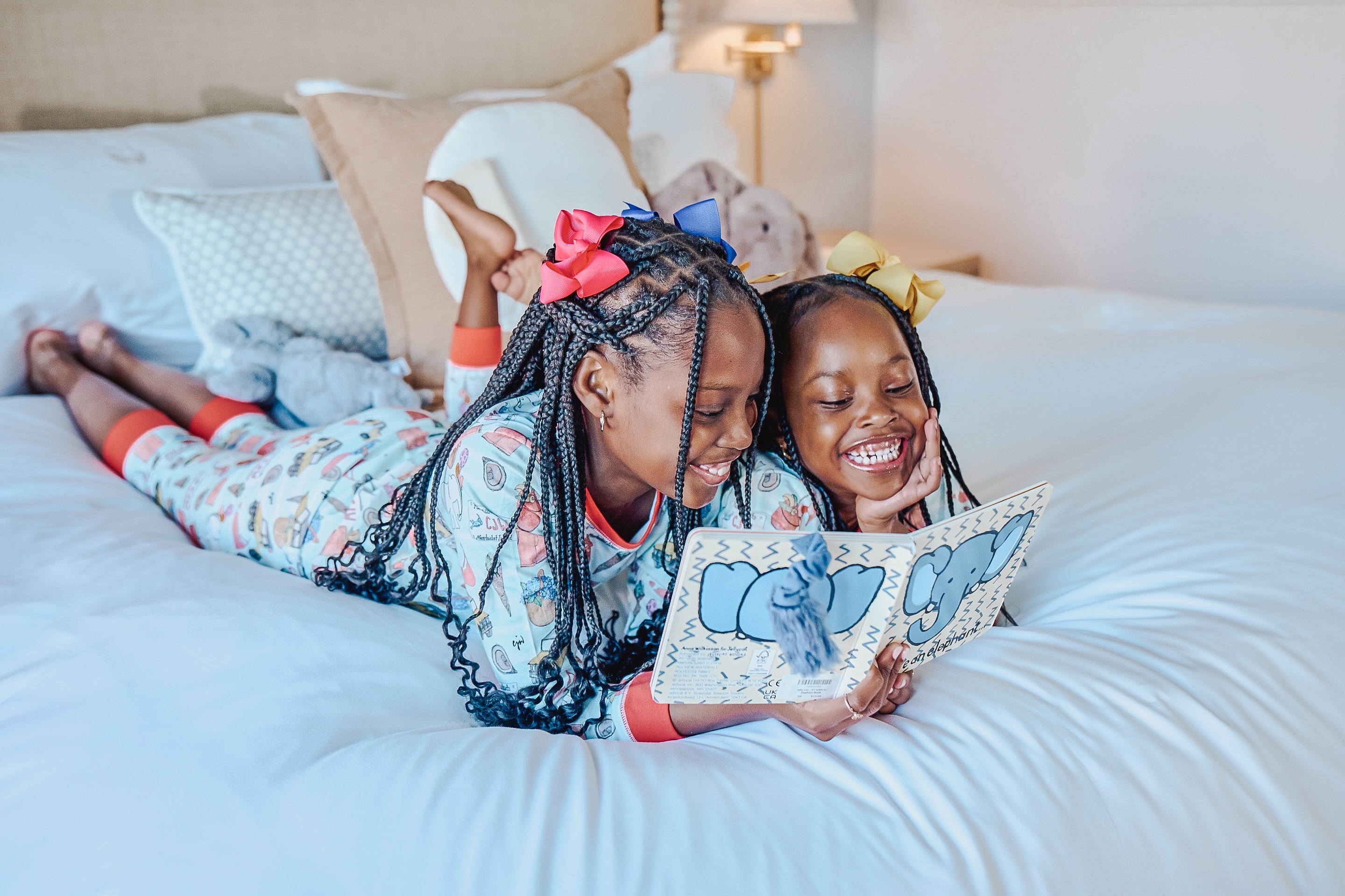 Two young girls with braided hair in pajamas lying on a bed reading a book.