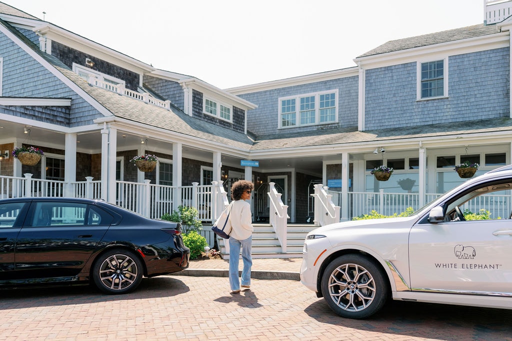 A blue-gray shingled building with white trim and a large porch, with a person and two cars in the foreground.