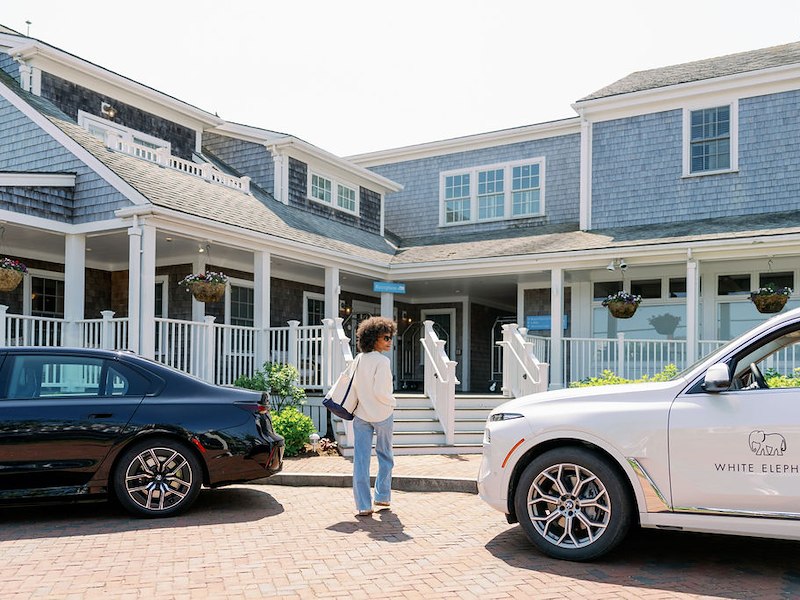 A blue-gray shingled building with white trim and a large porch, with a person and two cars in the foreground.