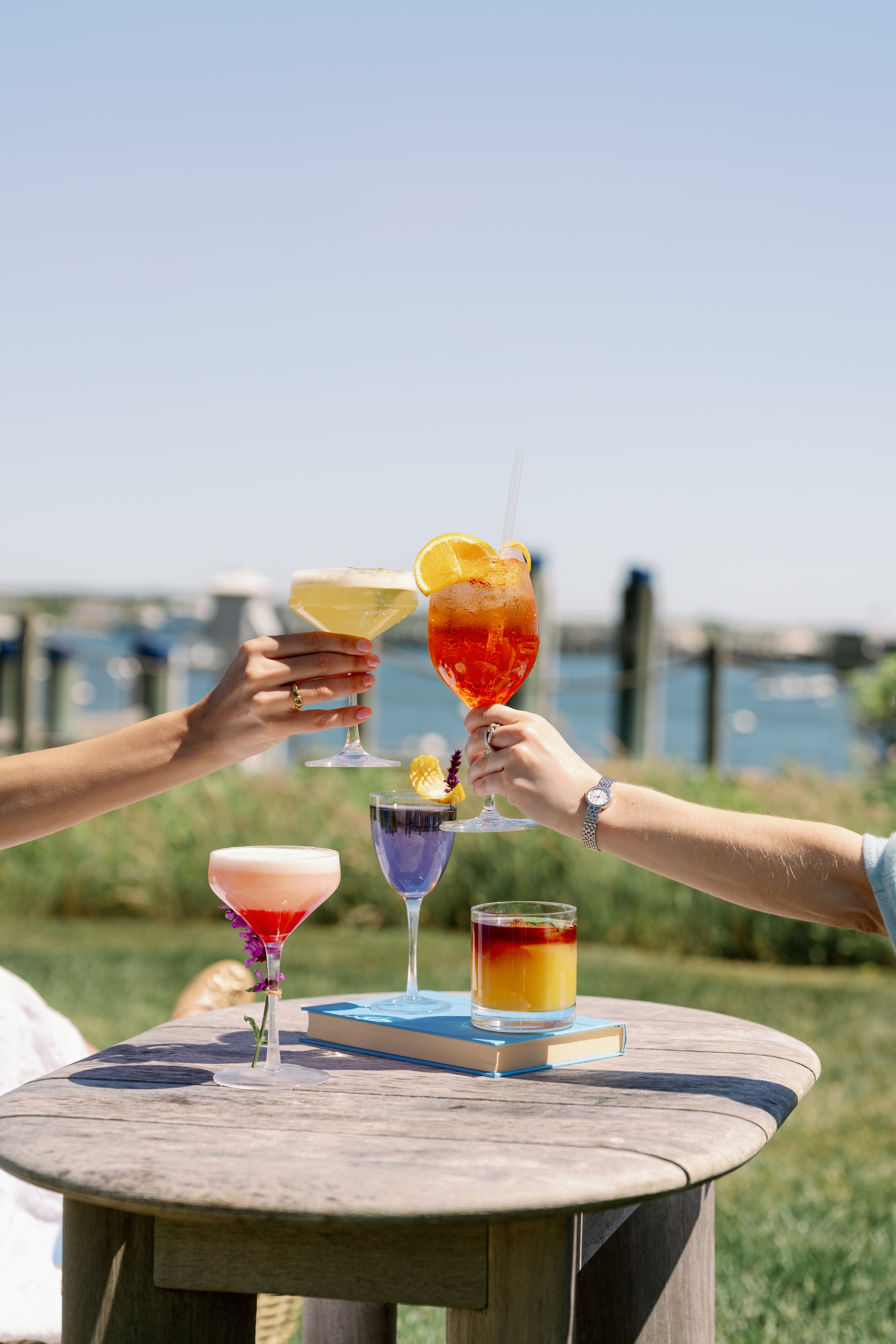 A table with colorful drinks and a book, with two people's arms reaching for the glasses.
