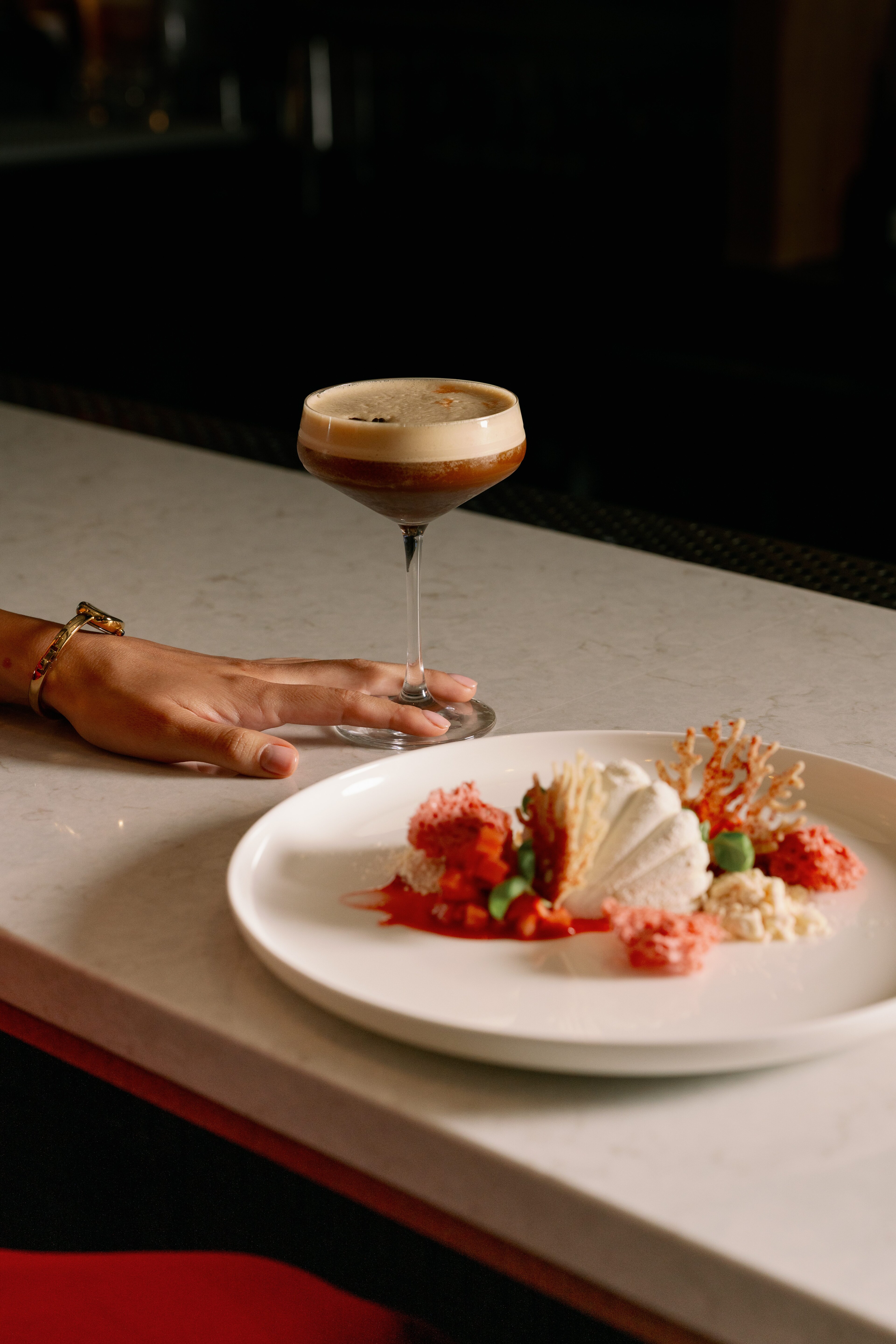 A hand rests on a bar next to a plate with food and a cocktail.