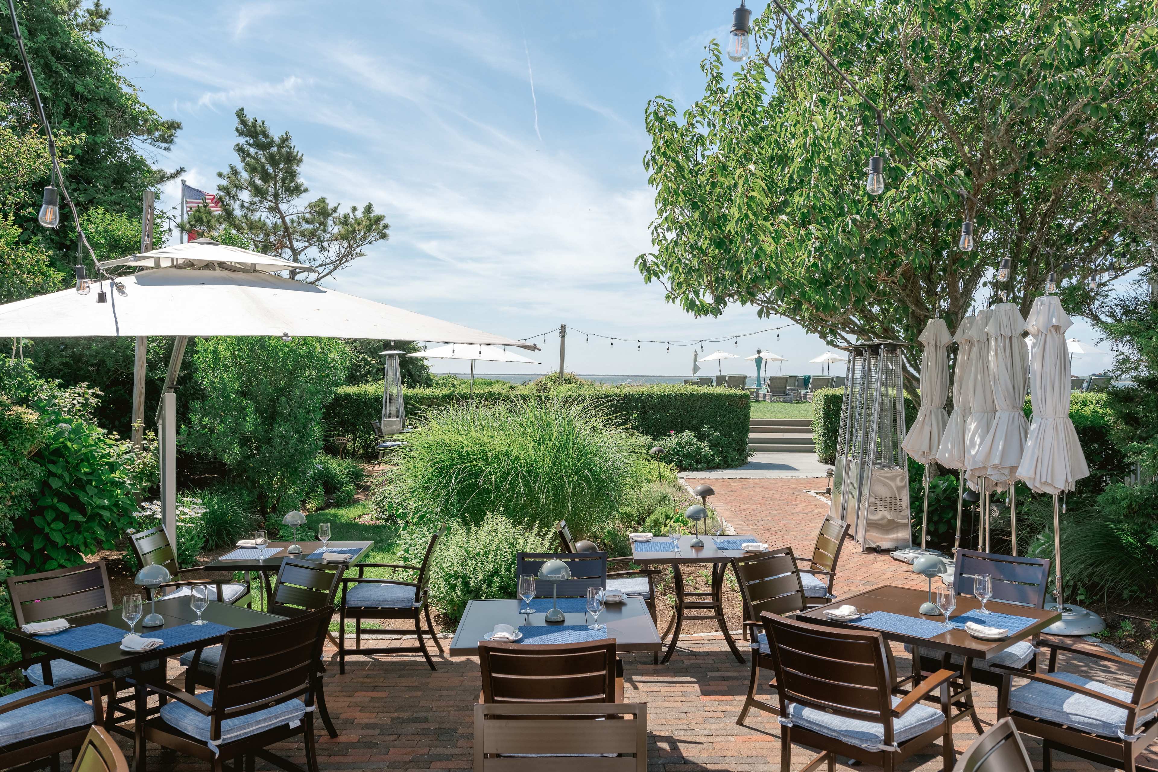 A patio dining area with tables and chairs set for meals, surrounded by lush greenery and trees, with string lights and umbrellas providing ambiance.