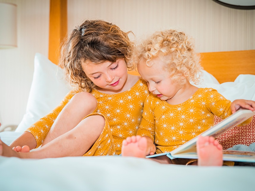 Two young girls in yellow shirts sitting on a bed, reading a book.