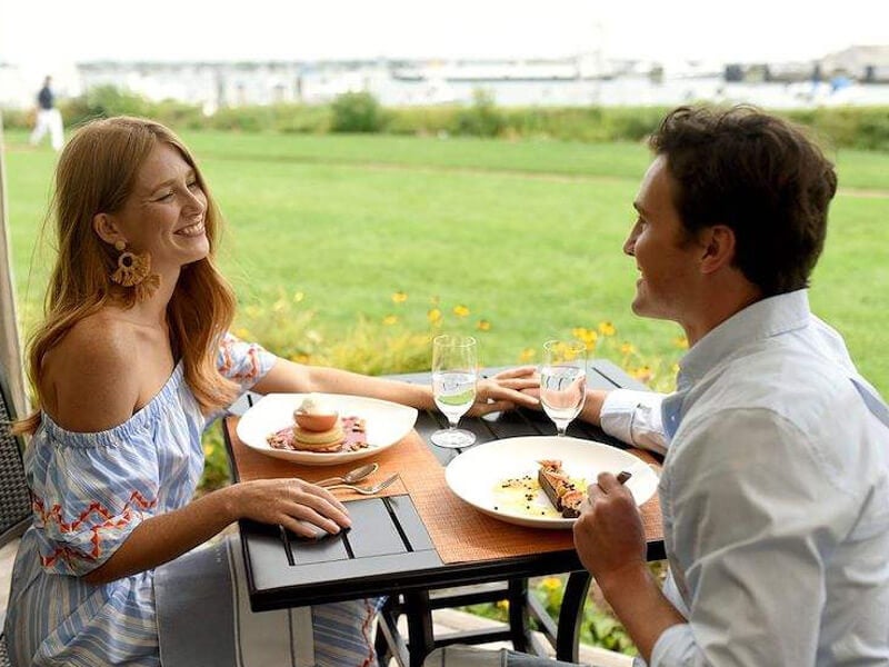 A couple enjoys a meal at an outdoor dining table with a view of a grassy area.