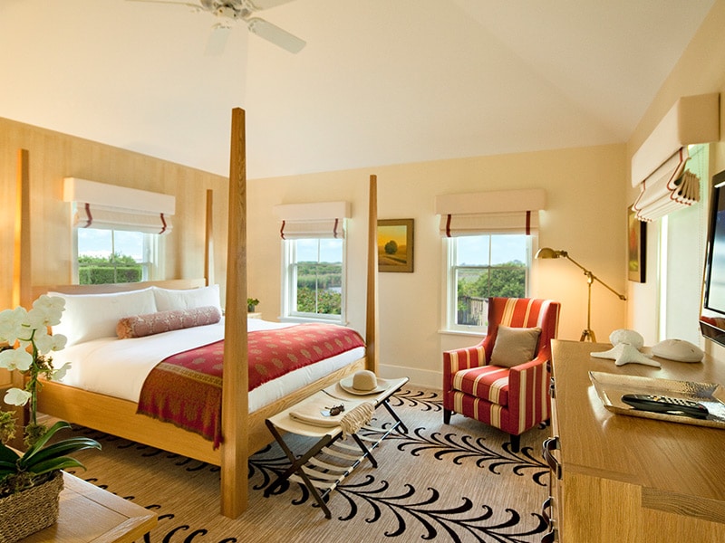 A wide-angle view of a bedroom with a four-poster bed, a red and white striped chair, a wooden dresser, and a ceiling fan.