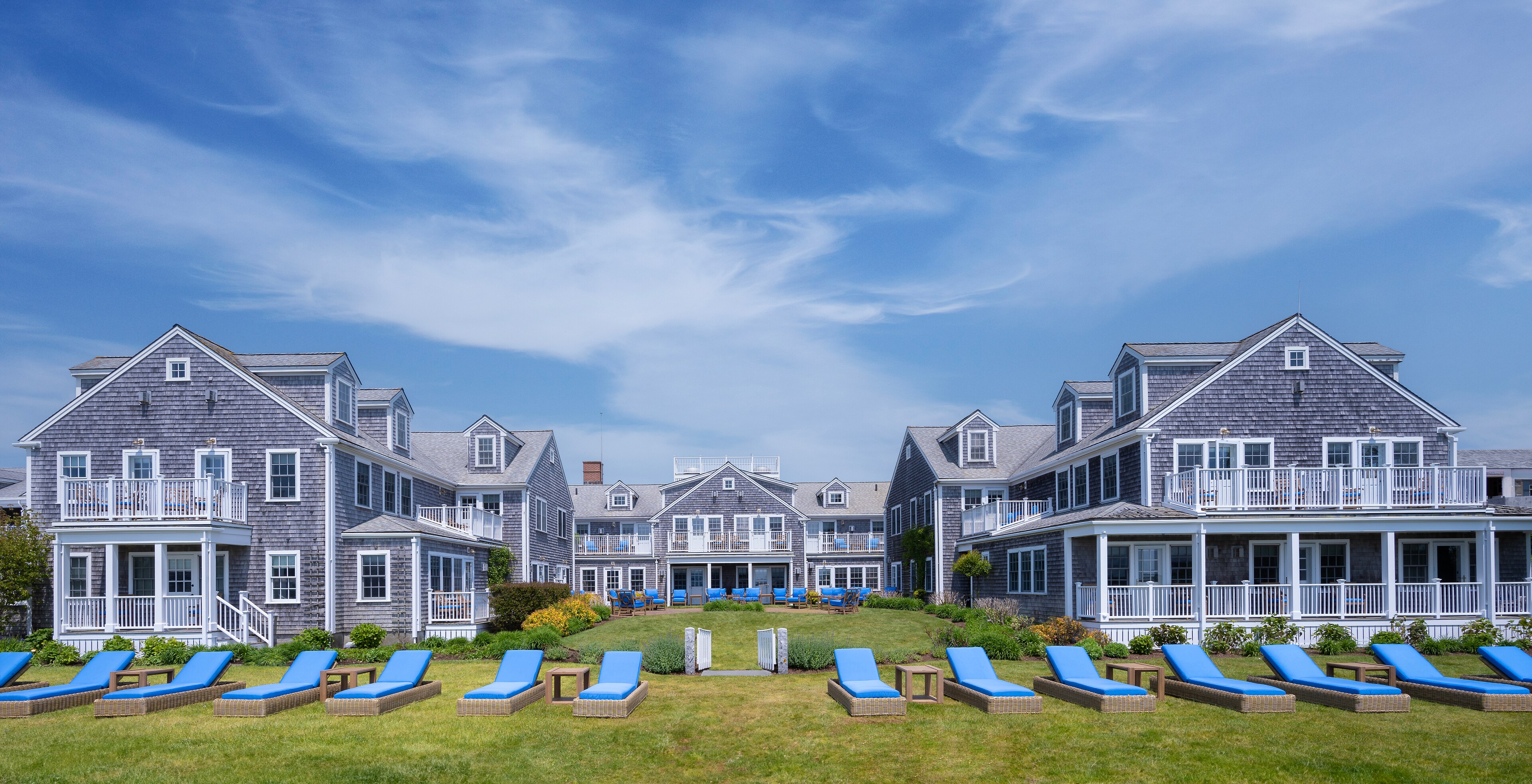 A large gray shingled building with white trim and balconies, with blue lounge chairs on the lawn in front.