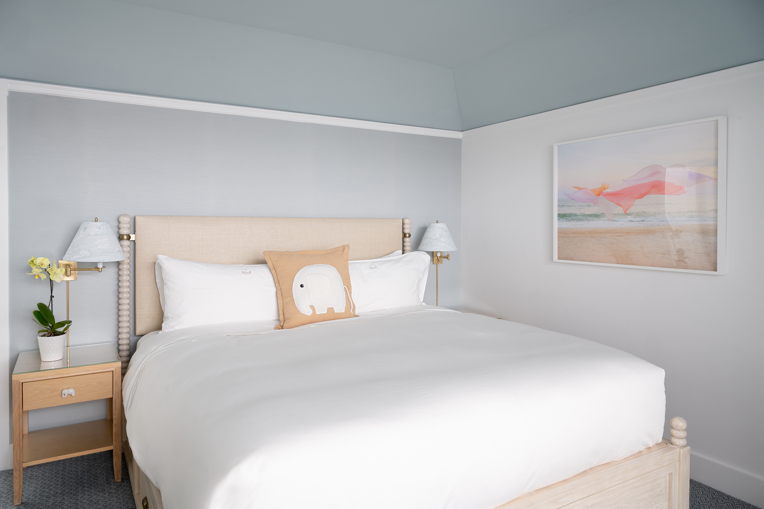 A neatly made bed with white bedding and a wooden frame, accompanied by two nightstands with lamps and a plant, in a room with light-colored walls and artwork.