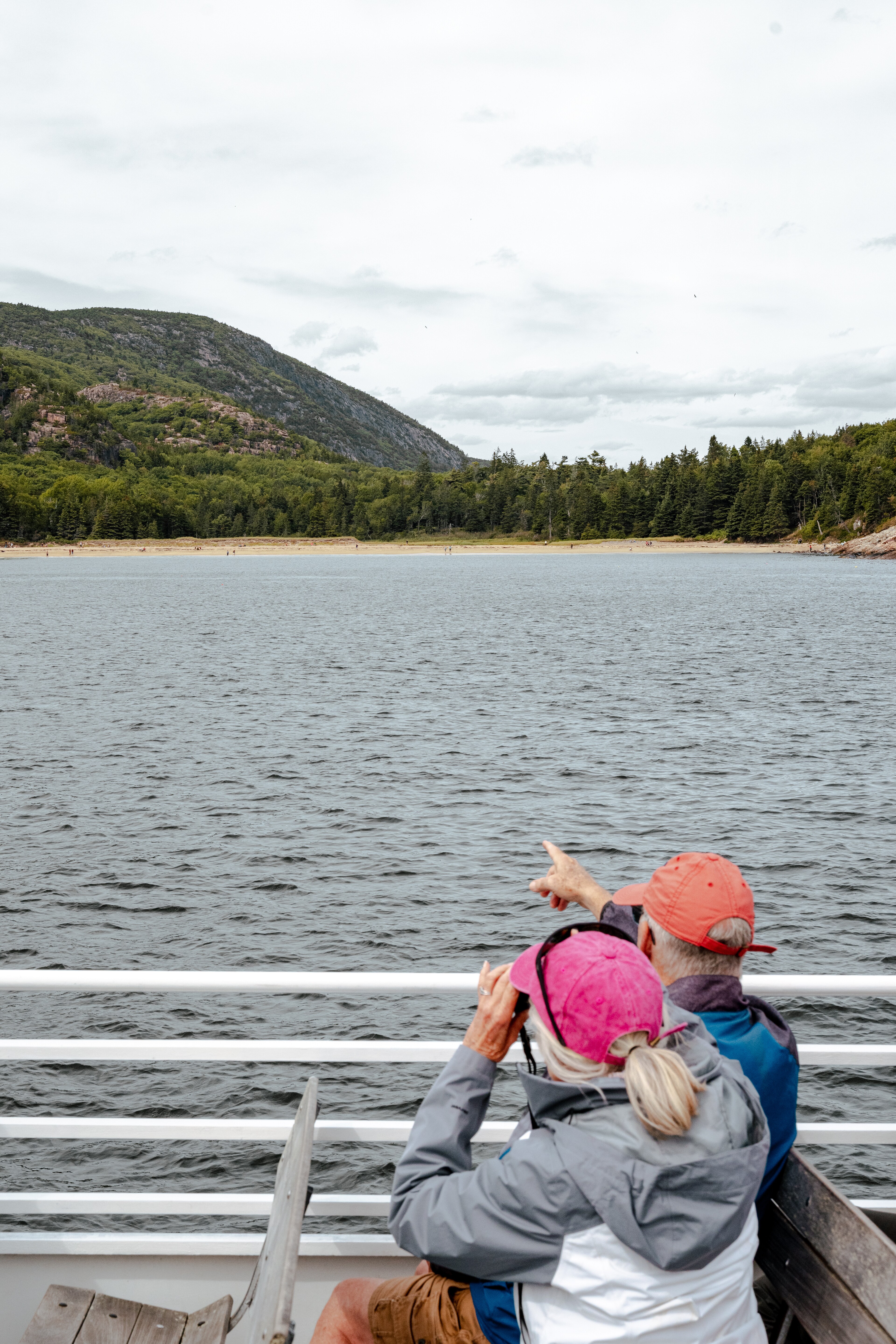 A couple wearing colorful jackets and caps sit on a ferry, looking out at a serene lake