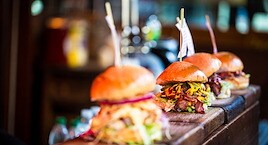 Four gourmet burgers with toothpicks are lined on a wooden counter