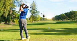 Golfer in blue shirt follows through on a swing, standing on a sunlit green fairway surrounded by trees