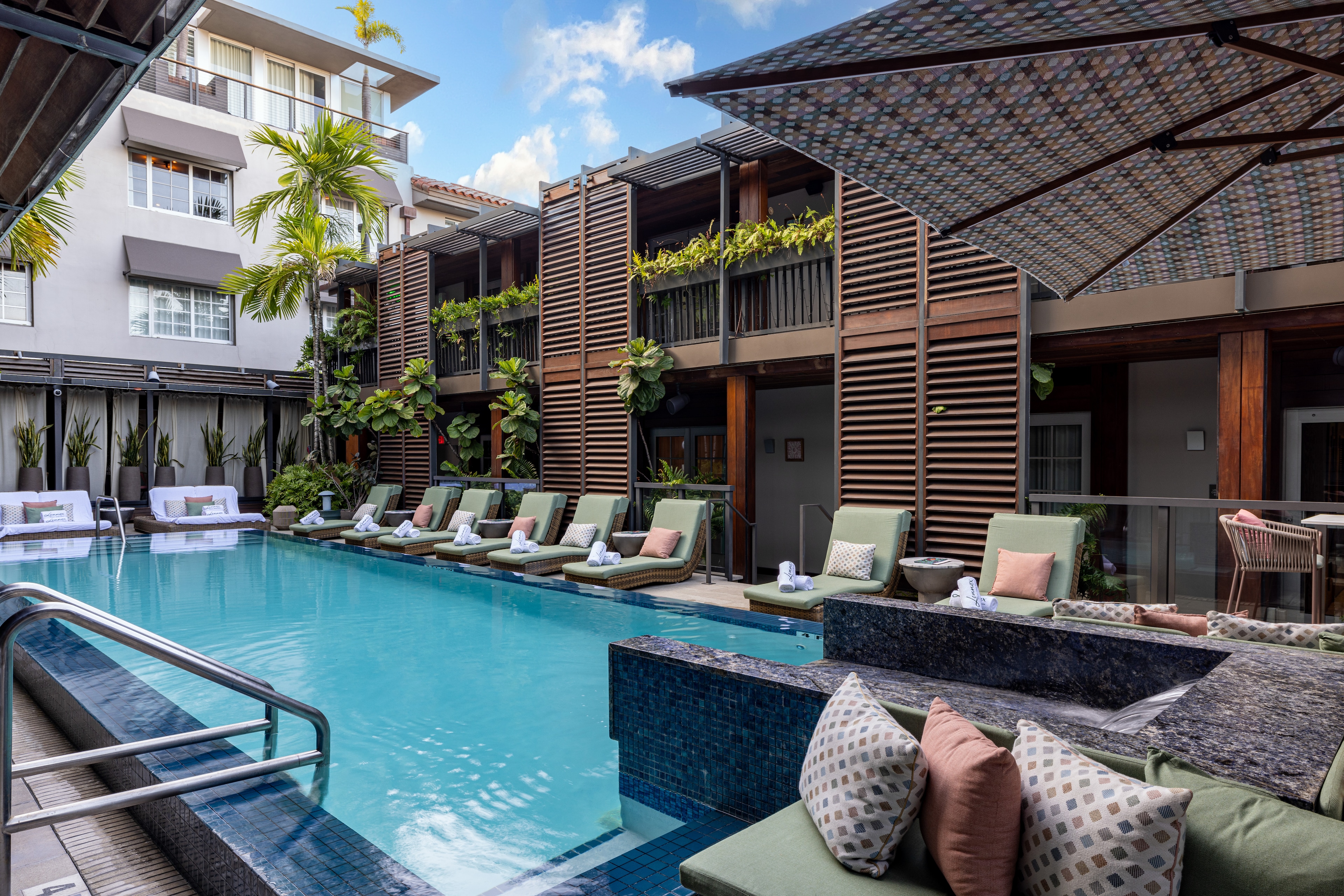A swimming pool with lounge chairs and umbrellas in front of a building with balconies and plants.