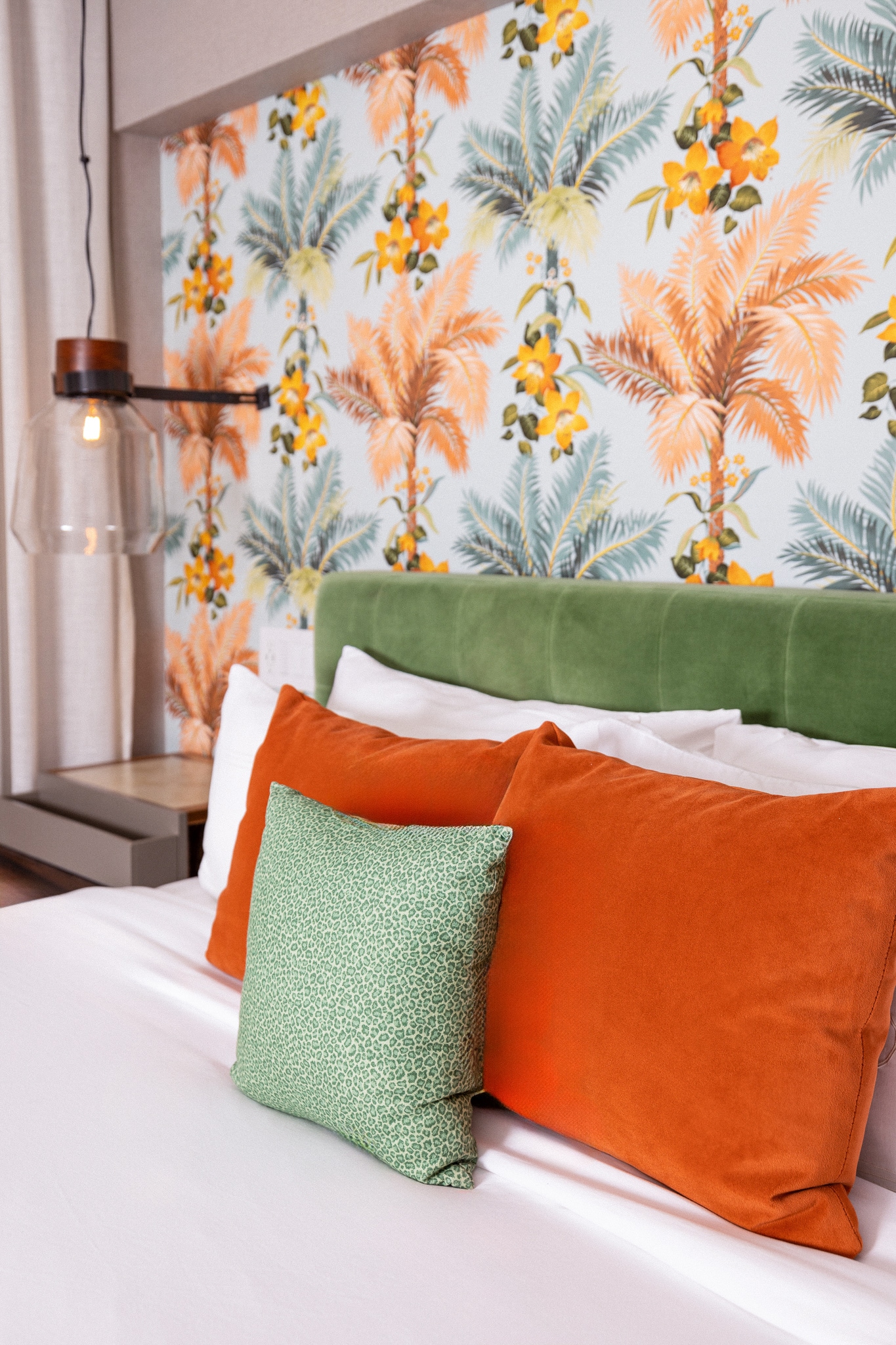 A close-up of a bed with a green headboard, orange pillows, and tropical wallpaper.