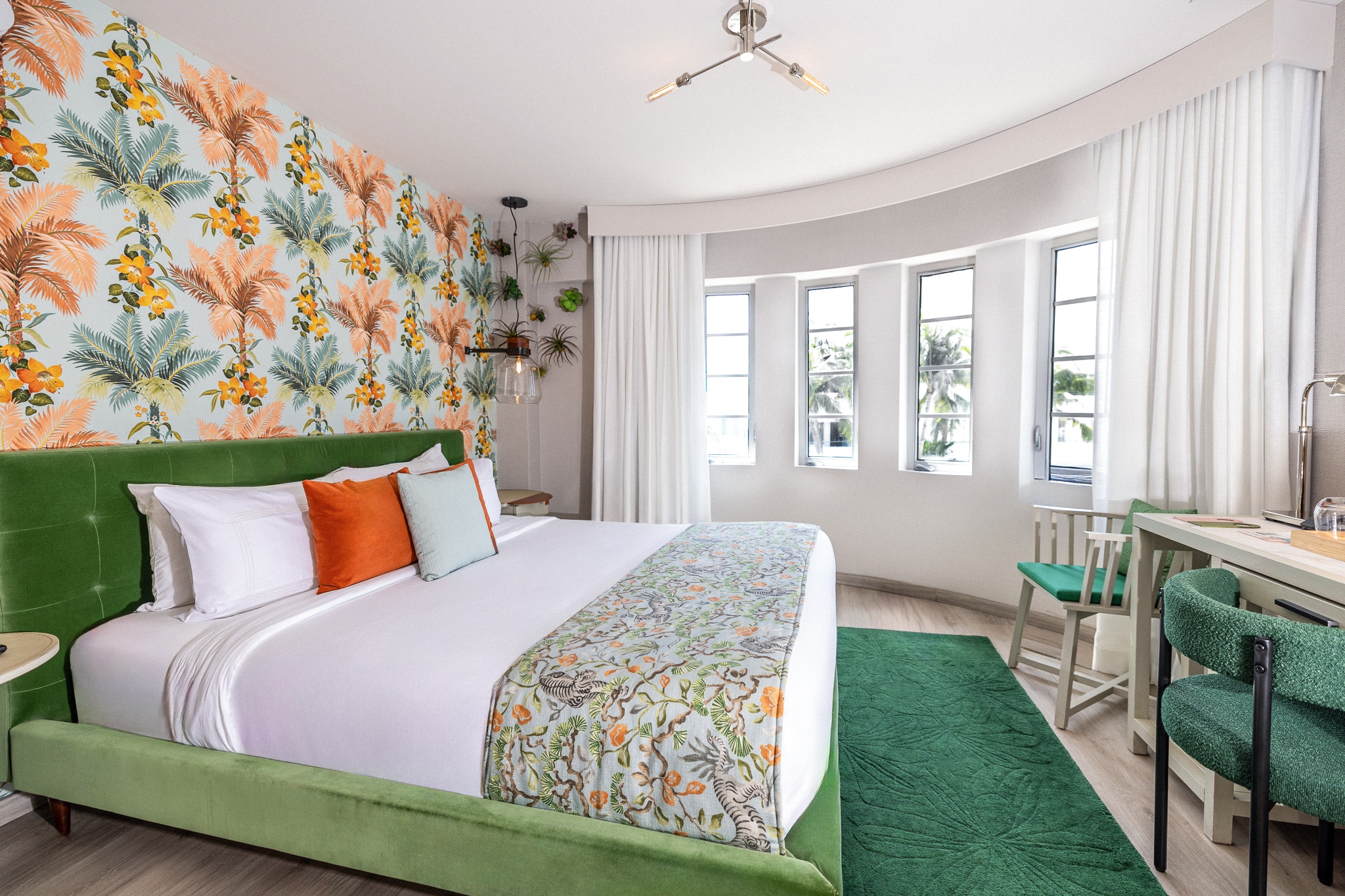 A brightly lit hotel room with a green bed, floral wallpaper, and a small dining area.