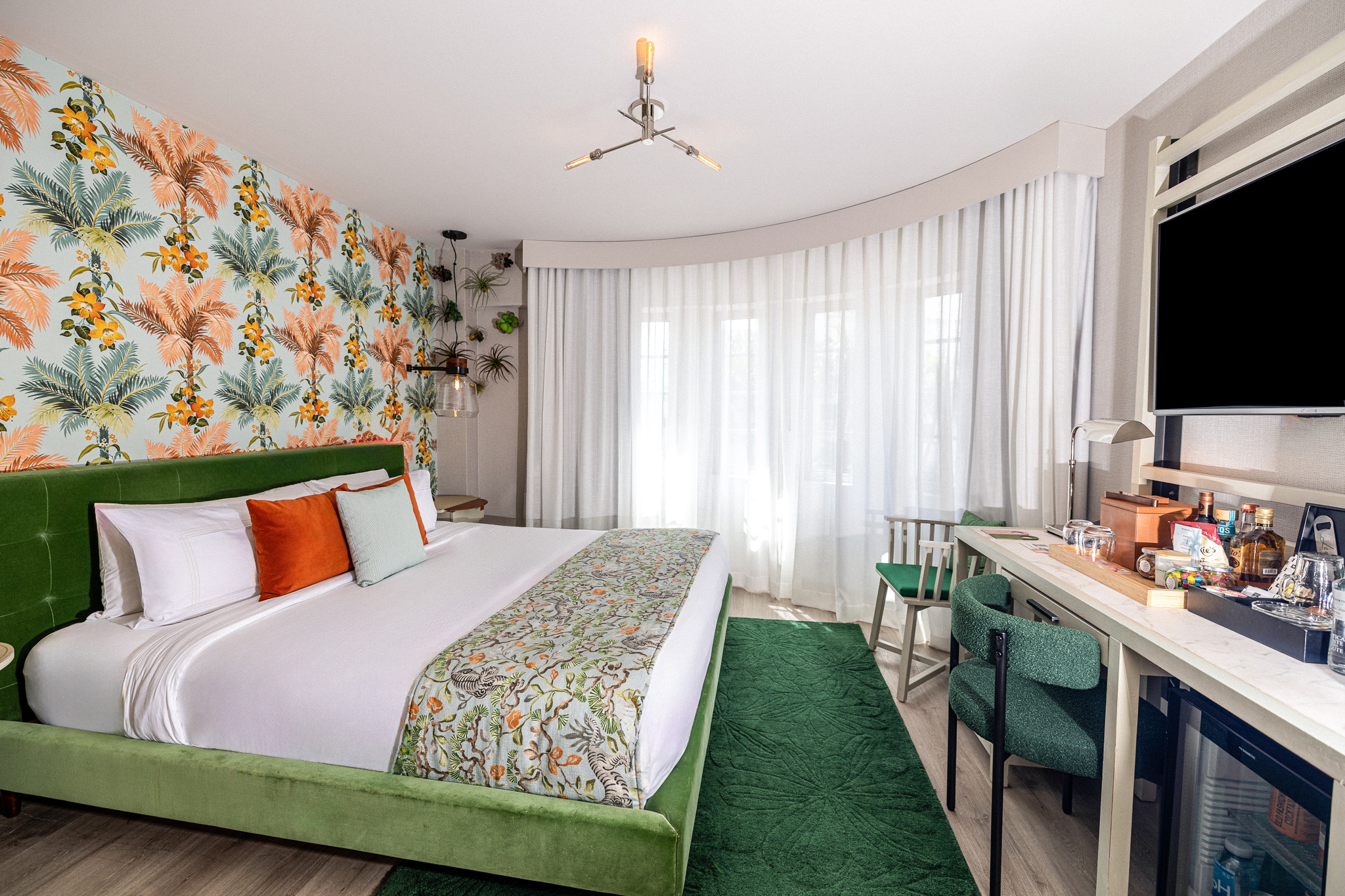 A hotel room with a green bed, desk, TV, and floral wallpaper
