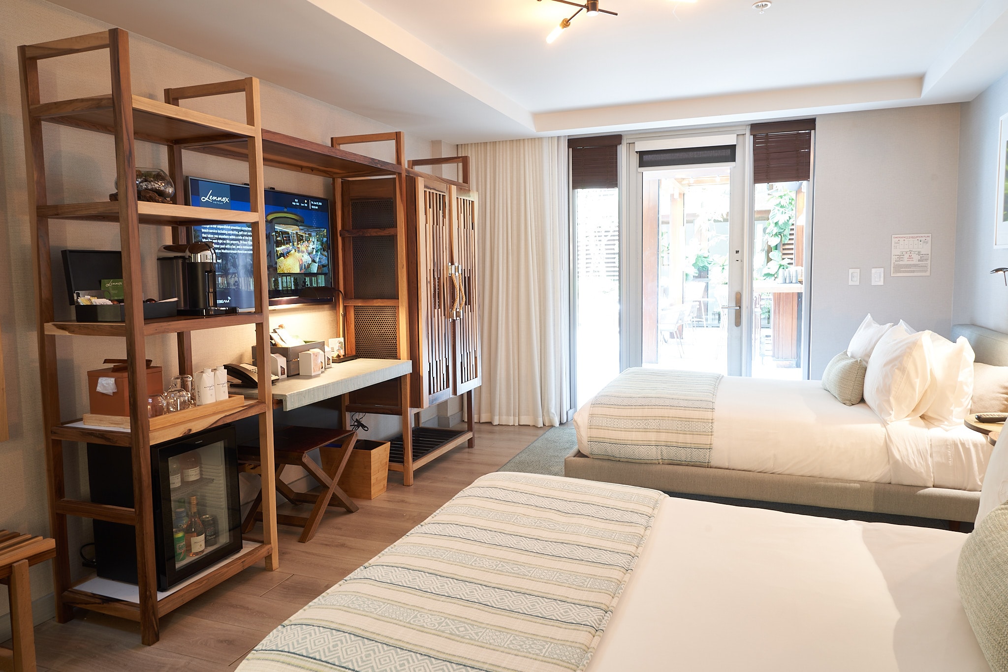 A hotel room with two beds, a desk, TV, mini_fridge, and shelving.
