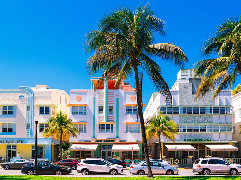 A colorful Art Deco building with cars parked in front and palm trees on the sidewalk.