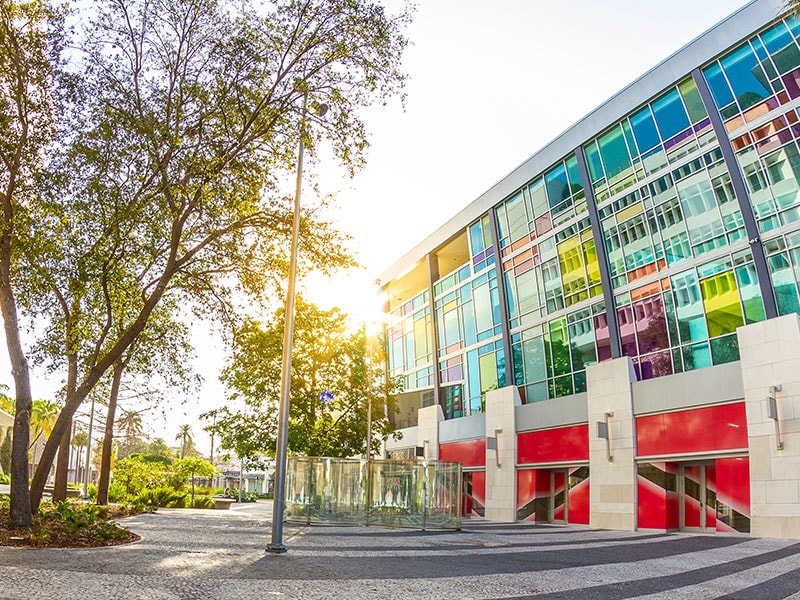 A multi-story building with a colorful glass facade and red accents, surrounded by trees and a streetlight.
