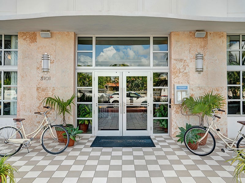 The main entrance of a building with glass doors, flanked by bicycles and plants, set in a stone facade with a tiled floor.