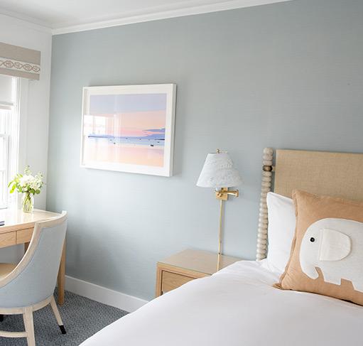 A neatly made bed with a white comforter and pillows, including one with an elephant design, in a room with light blue walls and a window.